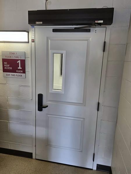 White stairwell door with lever handle, vision window and black overhead device on the header, beside a floor 1 stair sign