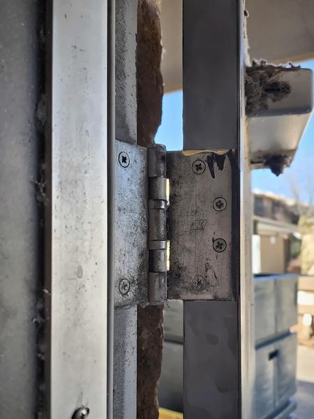 Close-up of a hollow metal door frame and hinge during repair, with an outdoor service yard behind